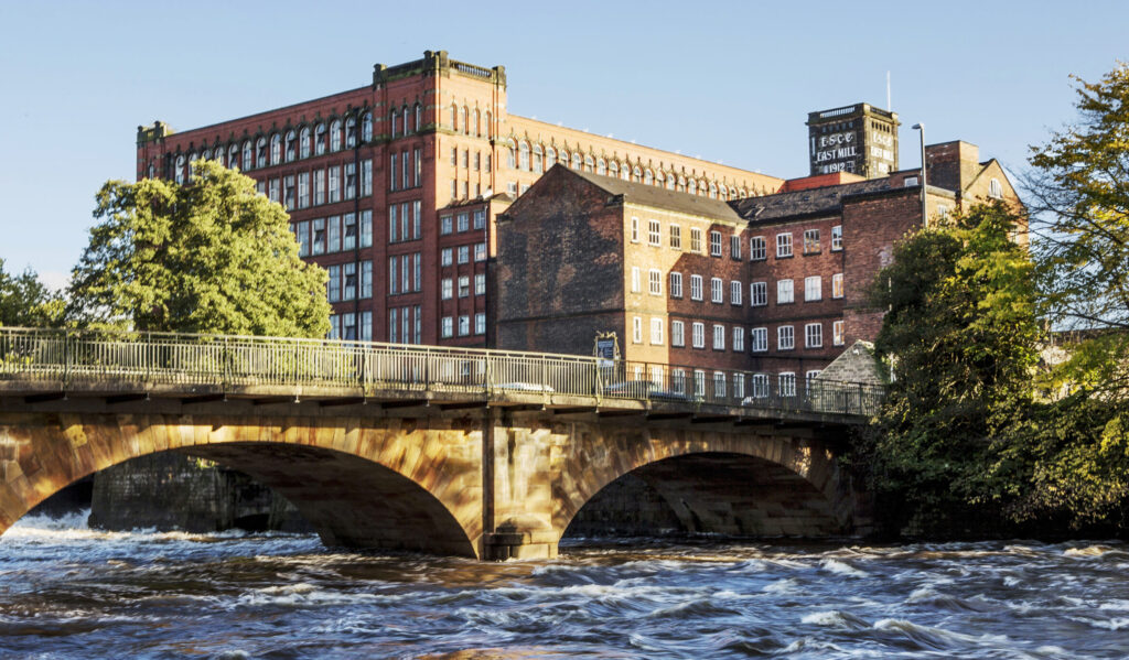 Belper East Mills, a large red brick building with multiple windows with the Belper North Mill building in front of it. The foreground is an arched road bridge with the River Derwent flowing underneath it.