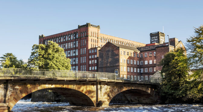 Belper East Mills, a large red brick building with multiple windows with the Belper North Mill building in front of it. The foreground is an arched road bridge with the River Derwent flowing underneath it.