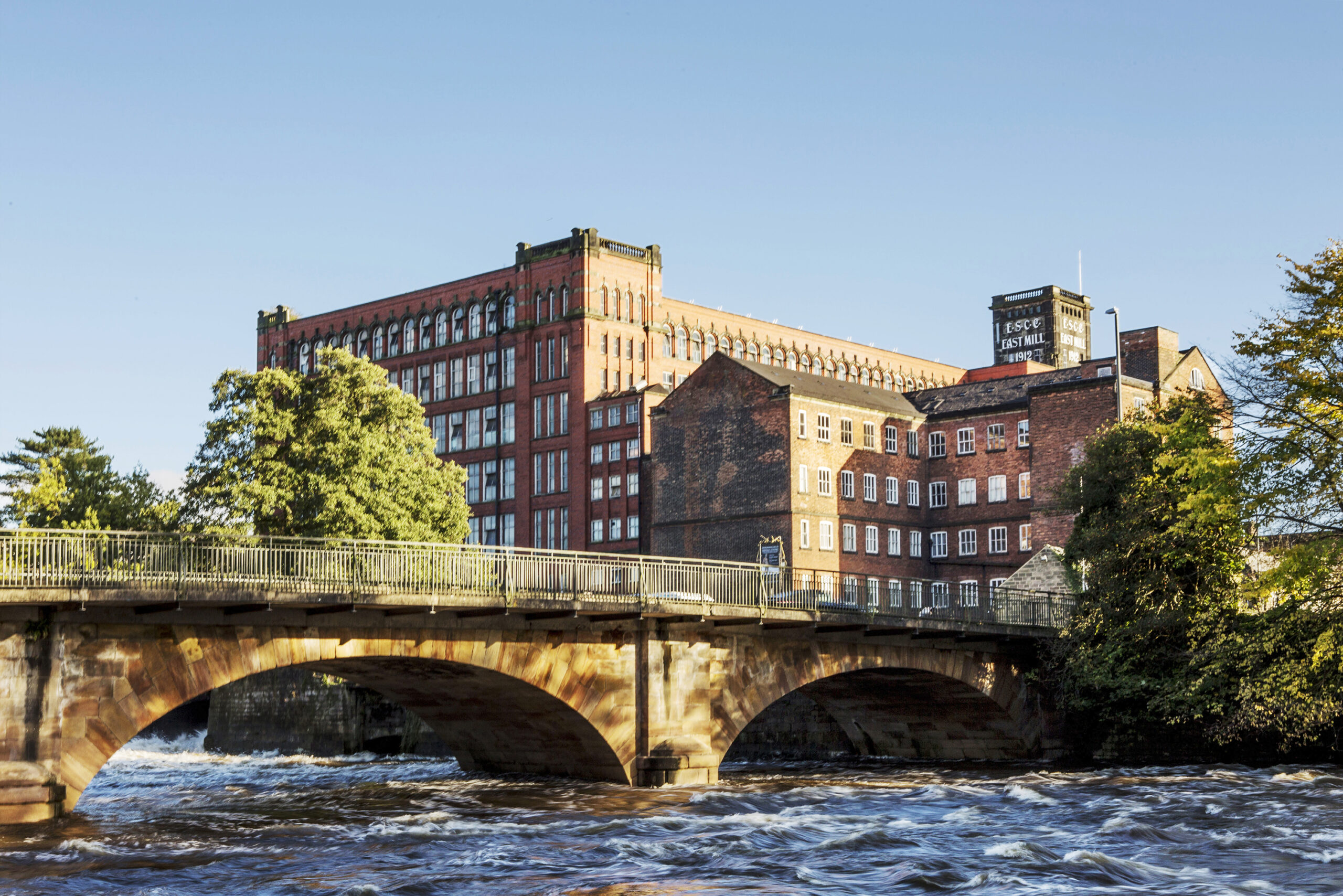 Belper East Mills, a large red brick building with multiple windows with the Belper North Mill building in front of it. The foreground is an arched road bridge with the River Derwent flowing underneath it.